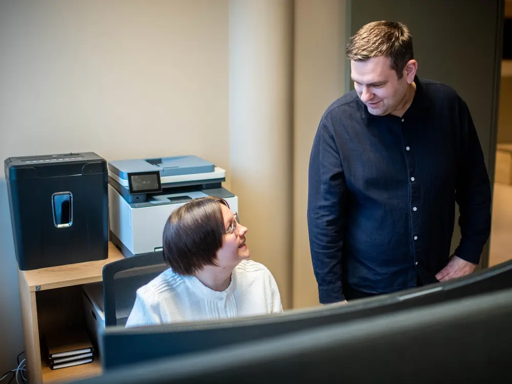 Kollega ved pulten smiler opp til en kollega som stikker innom